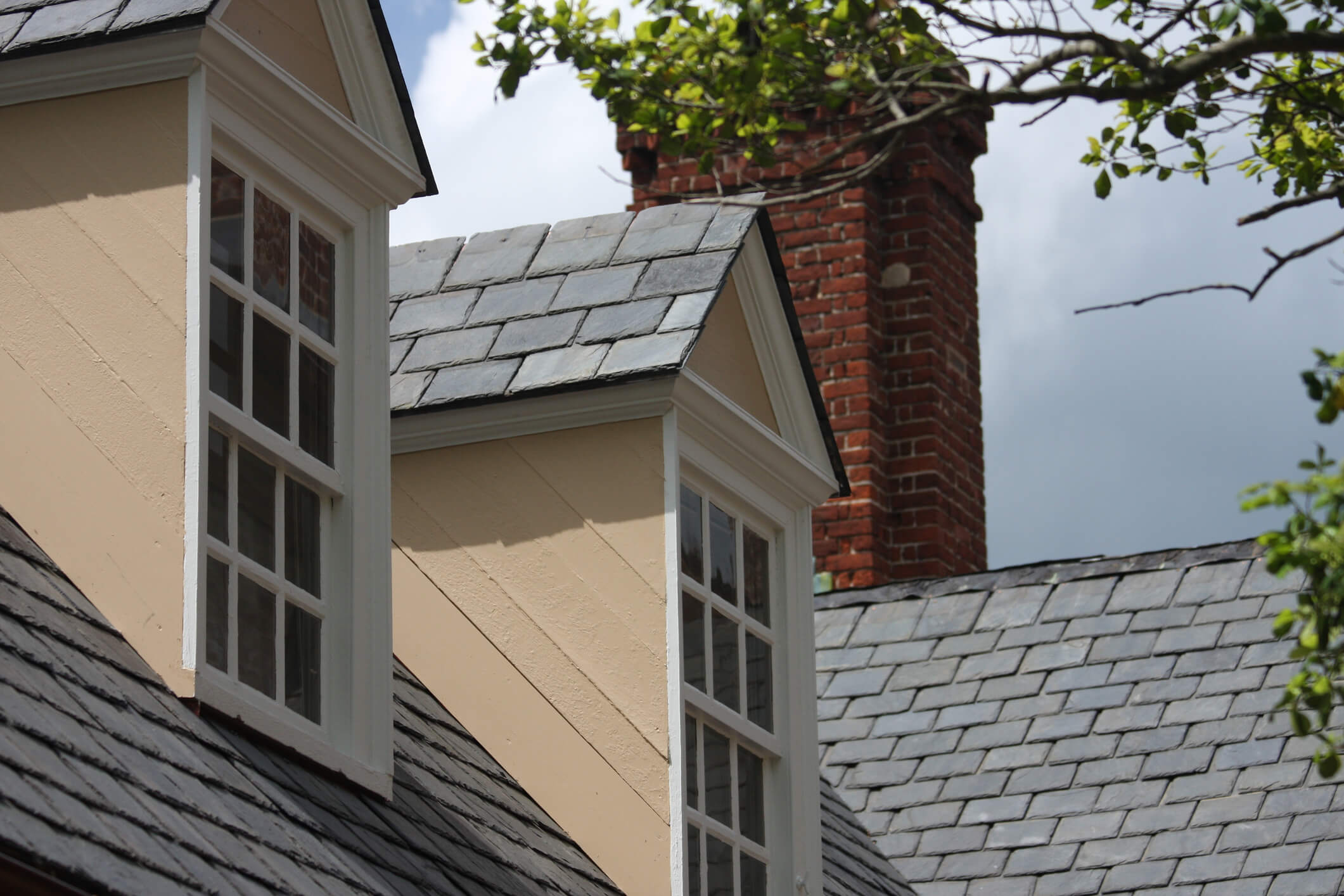 Roof of a house with light beige dormers and slate shingles. A red brick chimney rises in the background, framed by tree branches. Sky is partially cloudy.