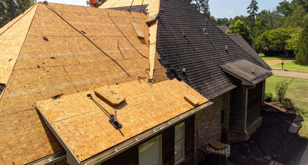 A house roof is partially covered with wooden sheathing, indicating ongoing construction or repair work by a roofer from Luling, LA. The left side remains unfinished, while the right boasts shingles. Scaffolding and tools are visible amid the lush greenery enveloping the house.