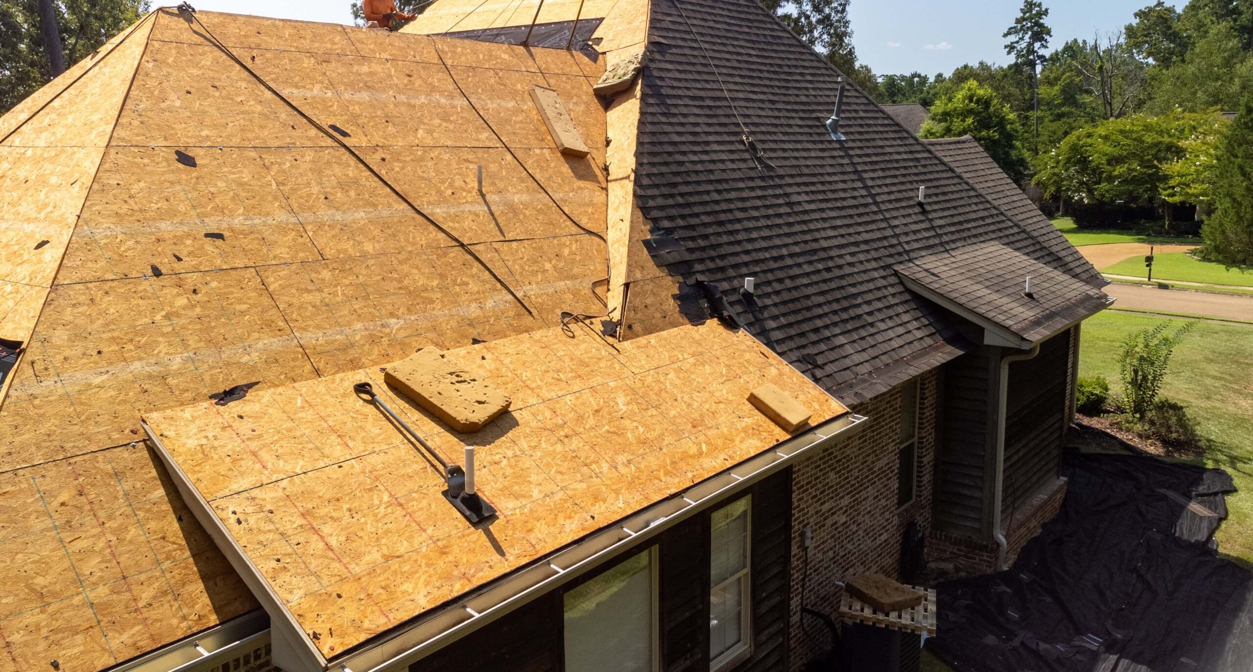 A house roof is partially covered with wooden sheathing, indicating ongoing construction or repair work by a roofer from Luling, LA. The left side remains unfinished, while the right boasts shingles. Scaffolding and tools are visible amid the lush greenery enveloping the house.