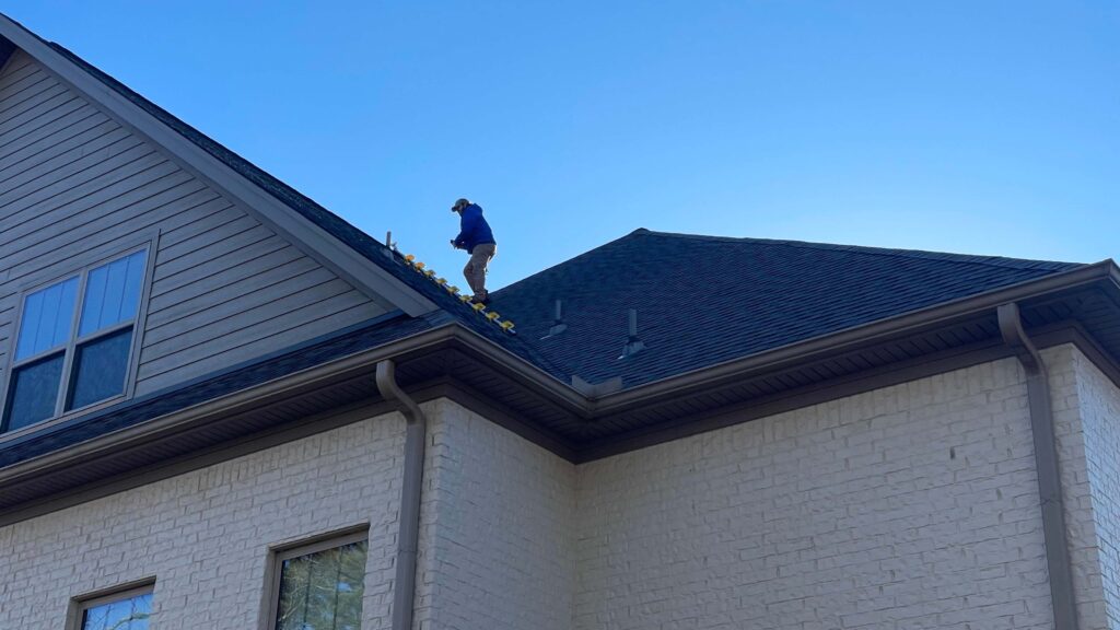 A roofer in River Oaks carefully clears leaves from the gutter of a two-story house. Their diligent work unfolds against a backdrop of light-colored brick walls, dark shingles, and a clear blue sky.