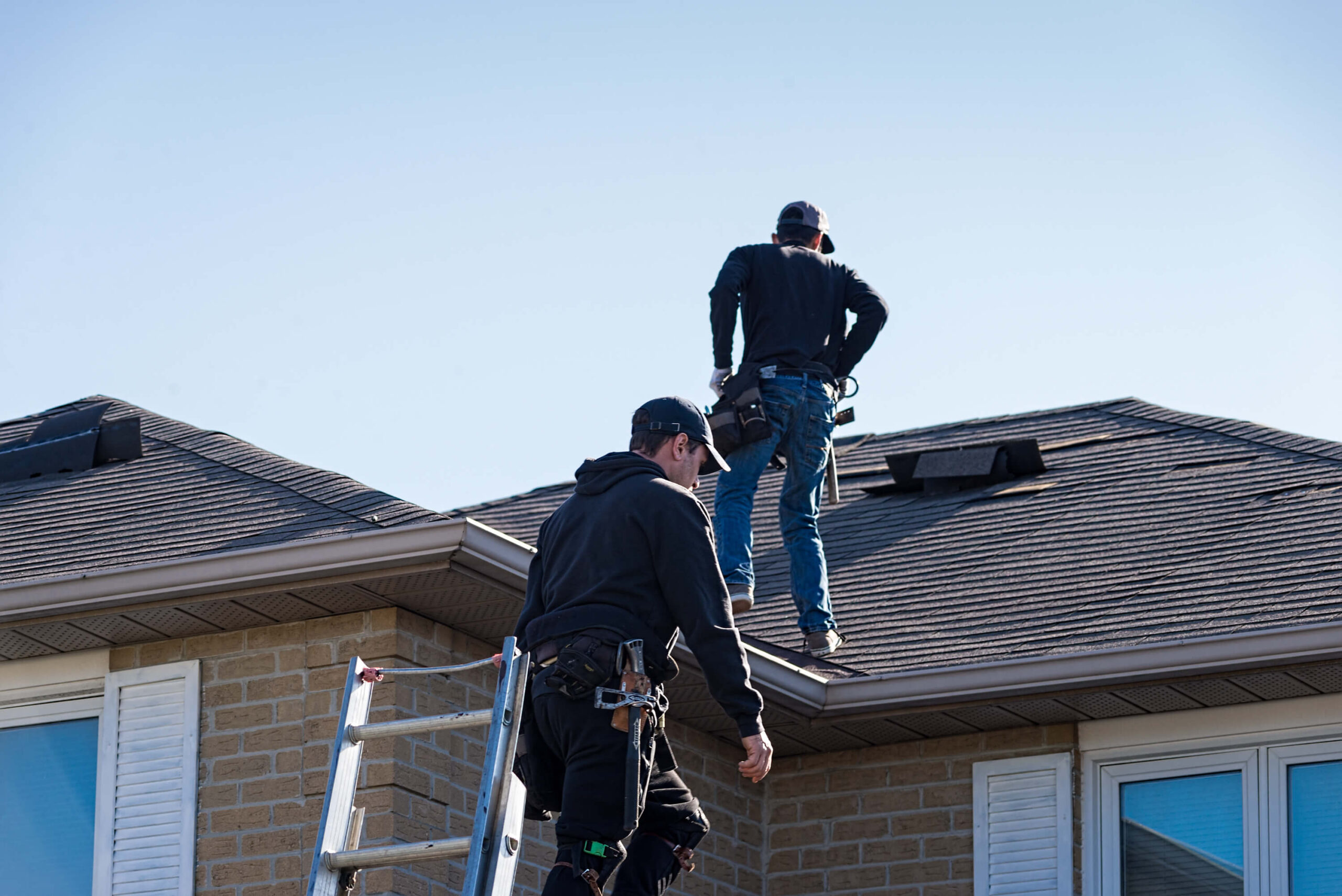Two roofers from St Rose, LA, dressed in black clothing, carefully inspect the roof of a beige brick house. One stands confidently on the roof while the other is perched on a ladder, both securely fastened in safety harnesses under the clear blue sky.