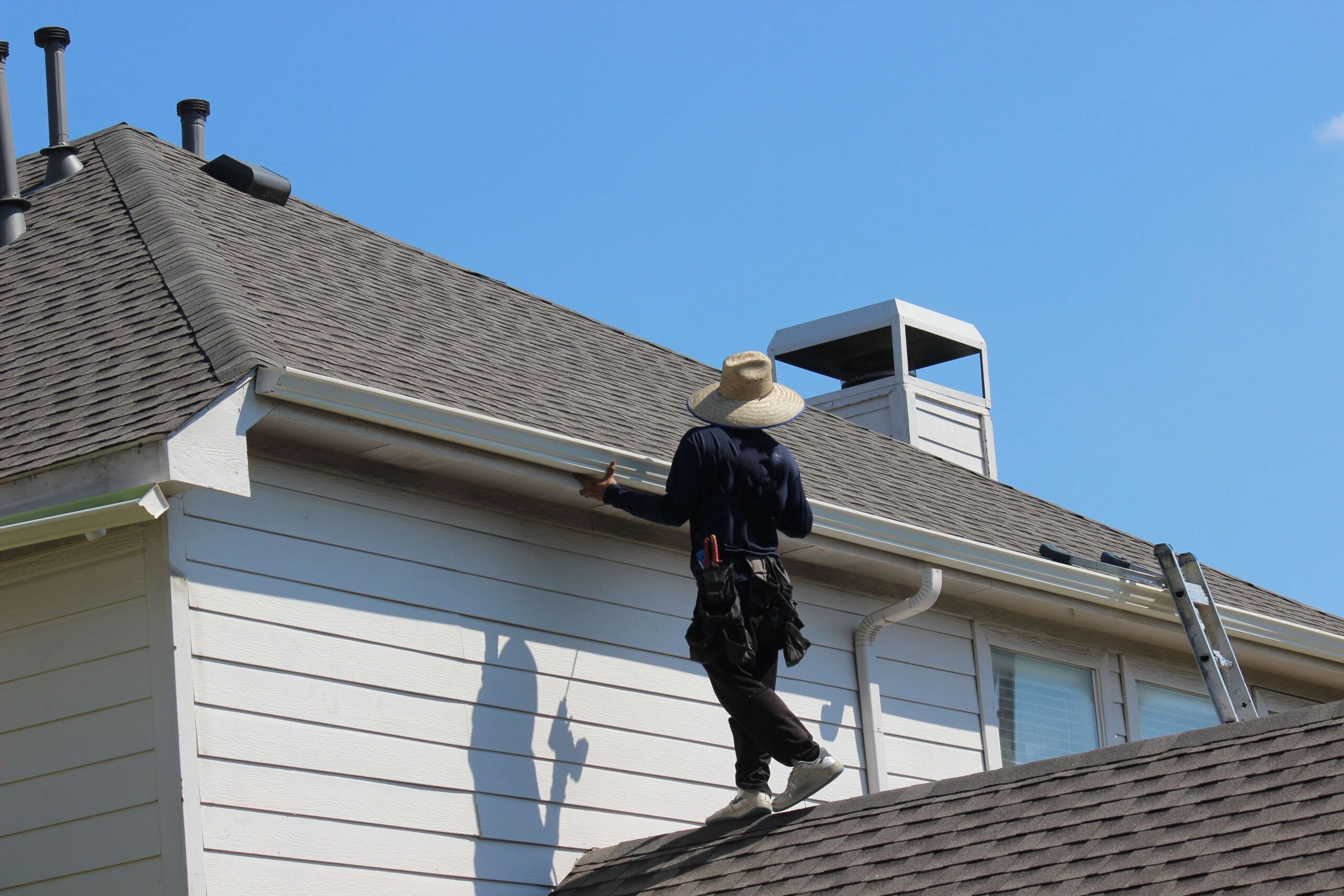 A roofer, wearing a wide-brimmed hat and tool belt, traverses the sloped roof of a house in the St. Rose Historic District. Near the edge where roof meets gutter, a ladder leans against the building under a clear blue sky.