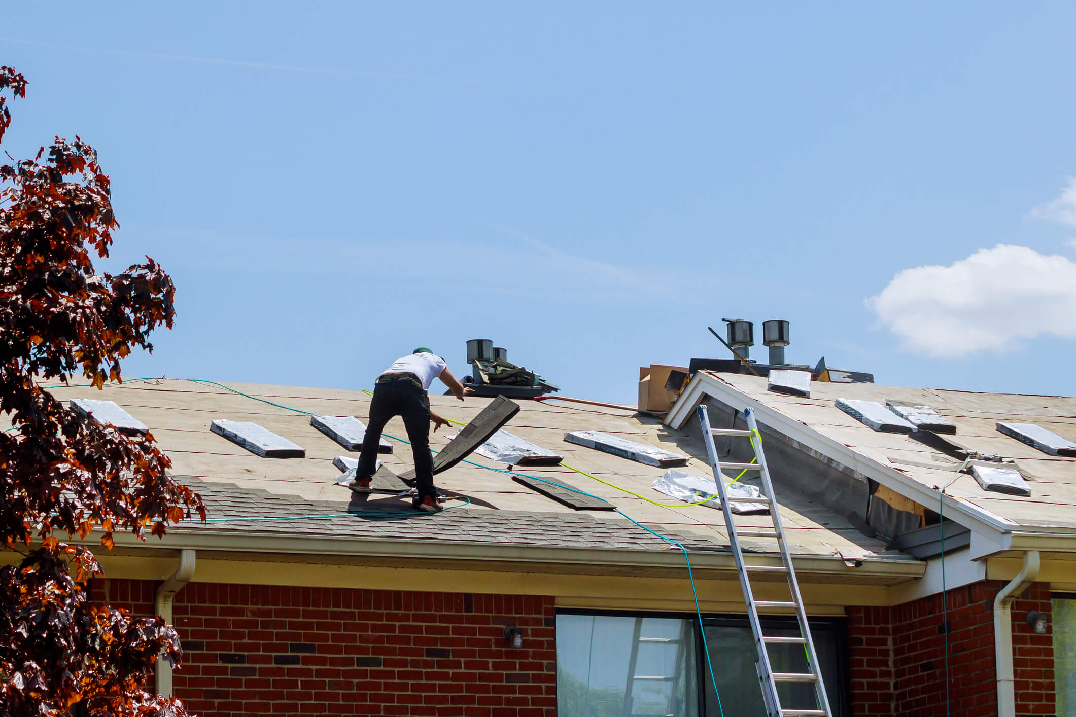 A worker in a white shirt and black pants is repairing a roof, surrounded by roofing materials. A metal ladder leans against the brick building, and the sky is clear and blue. Some trees are visible in the foreground.