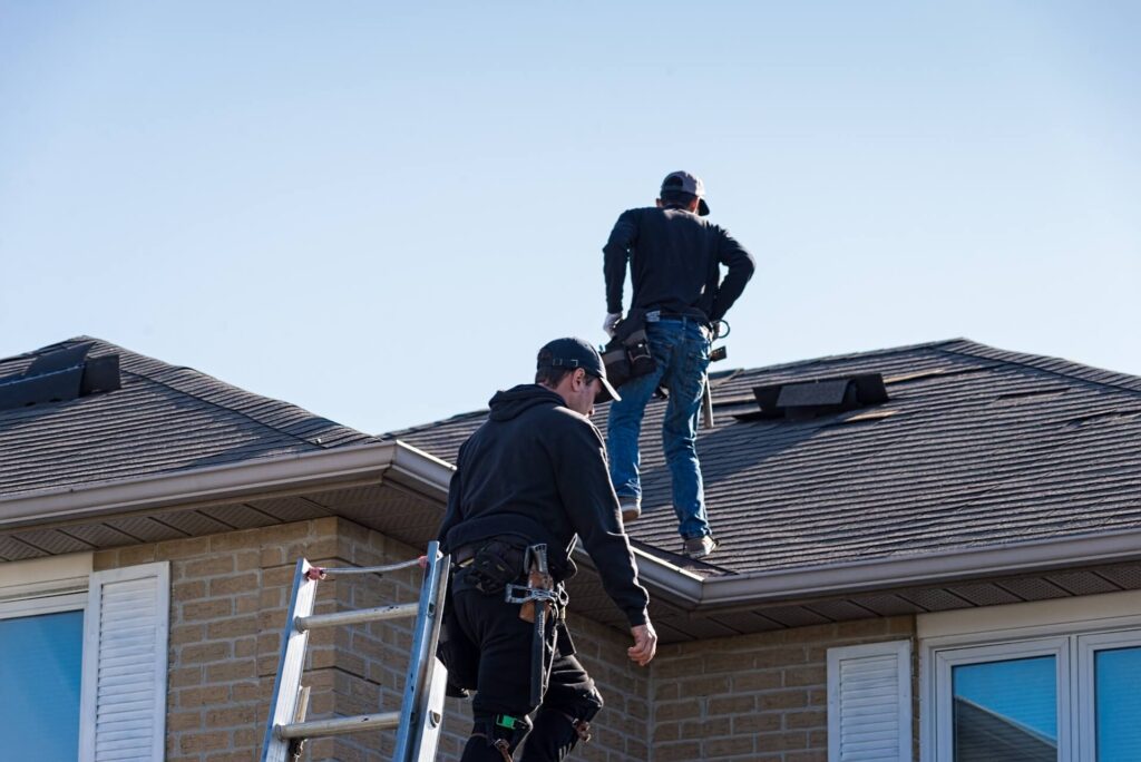 Two workers are standing on a roof, one near a ladder and the other further up. They are wearing casual clothes and caps, handling roof maintenance or repair tasks. The sky is clear, and the building is made of light-colored bricks.