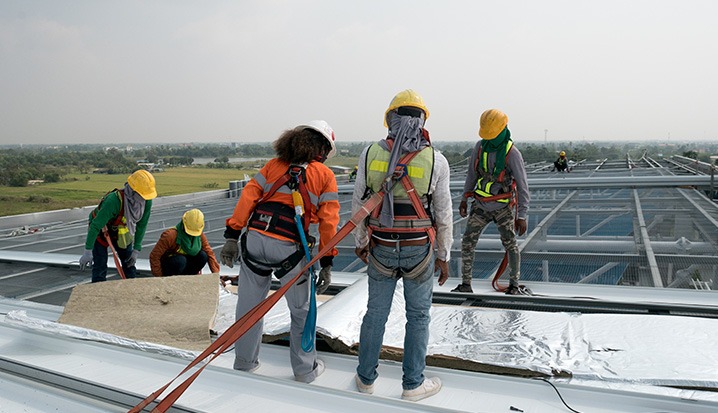 Five construction workers wearing safety gear and harnesses install insulation panels on a rooftop. A skilled New Orleans roofer leads the team, with open fields and trees visible in the background under a cloudy sky.