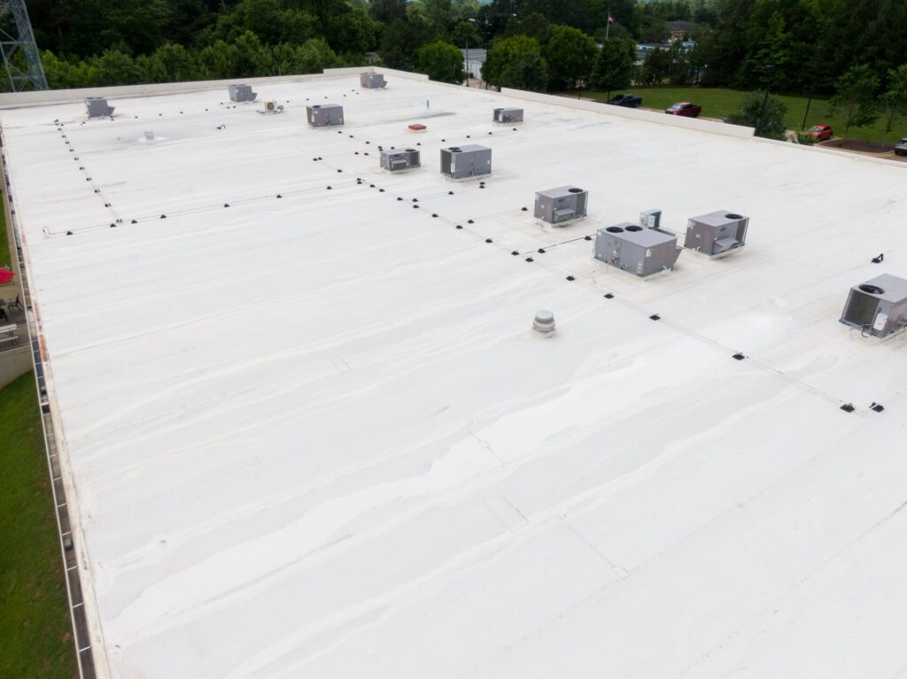 A large white commercial building roof with multiple HVAC units and vents, recently treated with Commercial Roof Coating in New Orleans, surrounded by trees and a few parked cars.