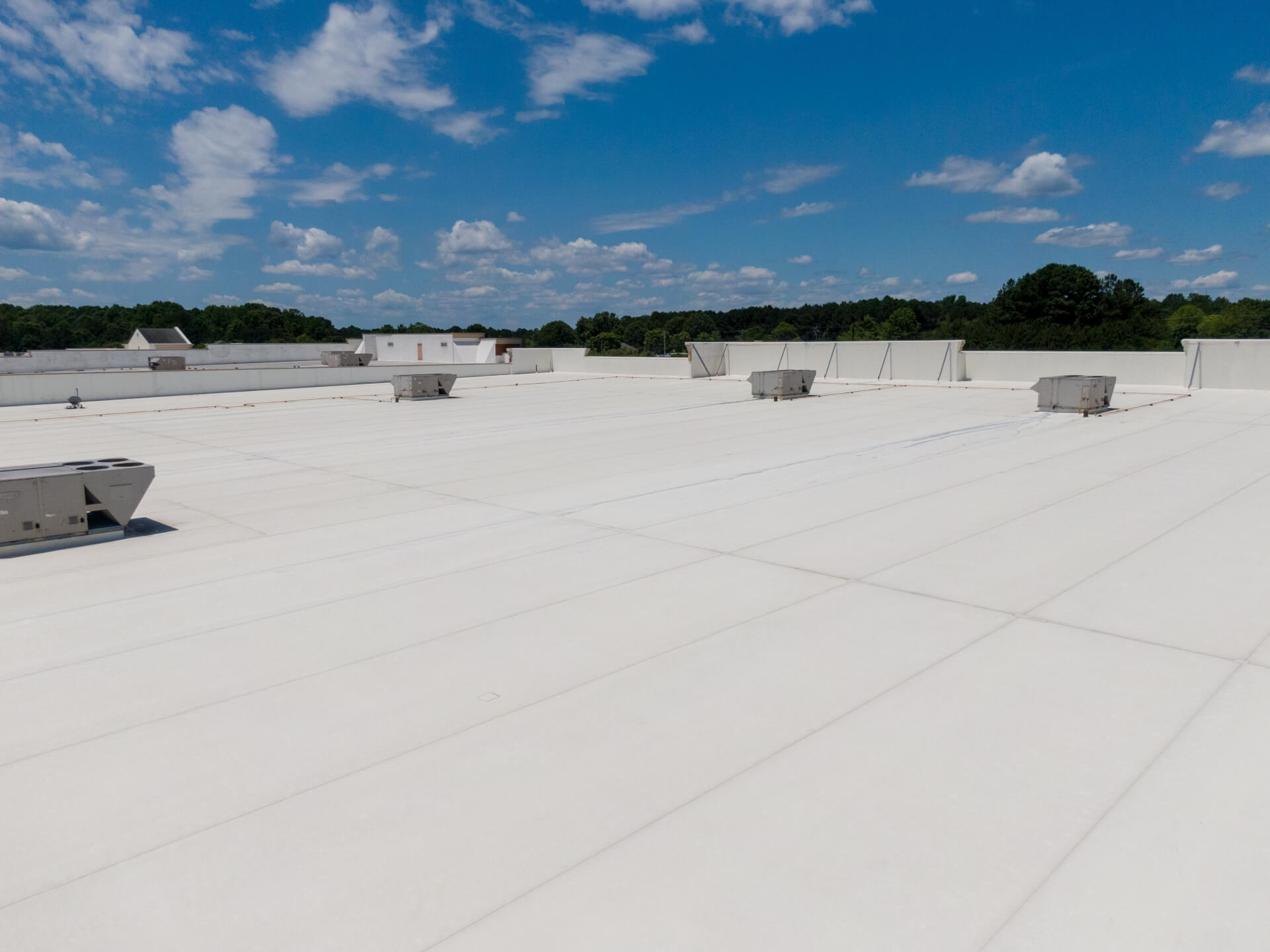 A large, flat white commercial rooftop with several HVAC units showcases the benefits of Commercial Roof Coating New Orleans, surrounded by a low wall, under a blue sky with scattered clouds and trees visible in the distance.