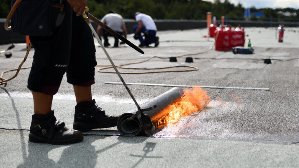 A worker uses a torch to install roofing material, melting and sealing it with flames. In the background, more workers handle roof repair New Orleans style on the flat rooftop, with equipment and tools visible nearby.