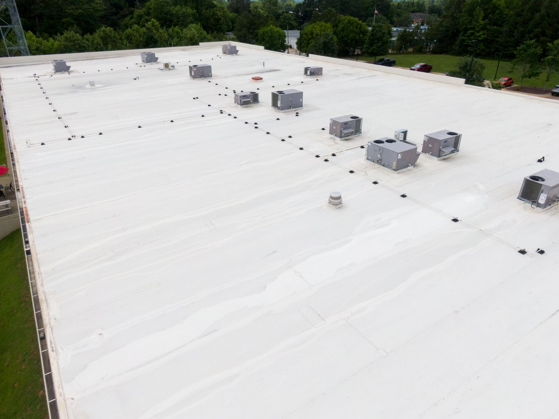 A large, flat white commercial rooftop with several HVAC units and vents showcases expert flat roofing New Orleans style, surrounded by green trees and a parking lot with a few cars in the background.