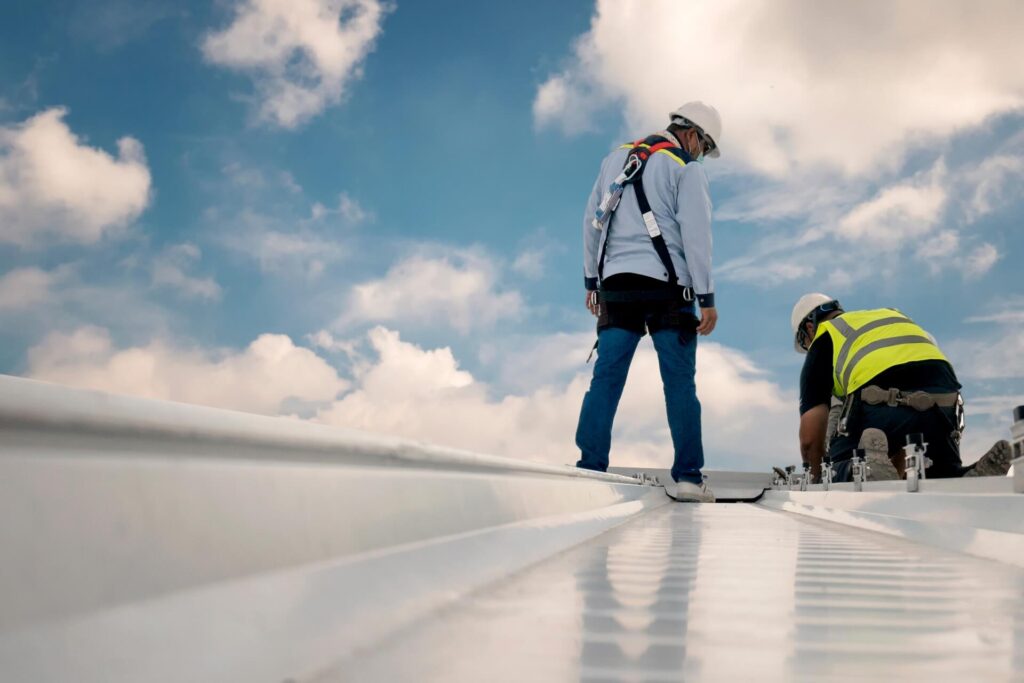 Two workers in safety gear and helmets inspect a metal rooftop under a blue sky with scattered clouds, possibly preparing for a Commercial Roof Coating New Orleans project. One stands while the other kneels, both focused on the rooftop surface.