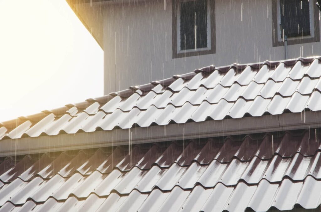 Rain falls on a metal roof of a house, with sunlight shining from the left side of the image. The wavy pattern highlights the quality of Metal Roofing New Orleans, while part of a window and wall can be seen in the background.