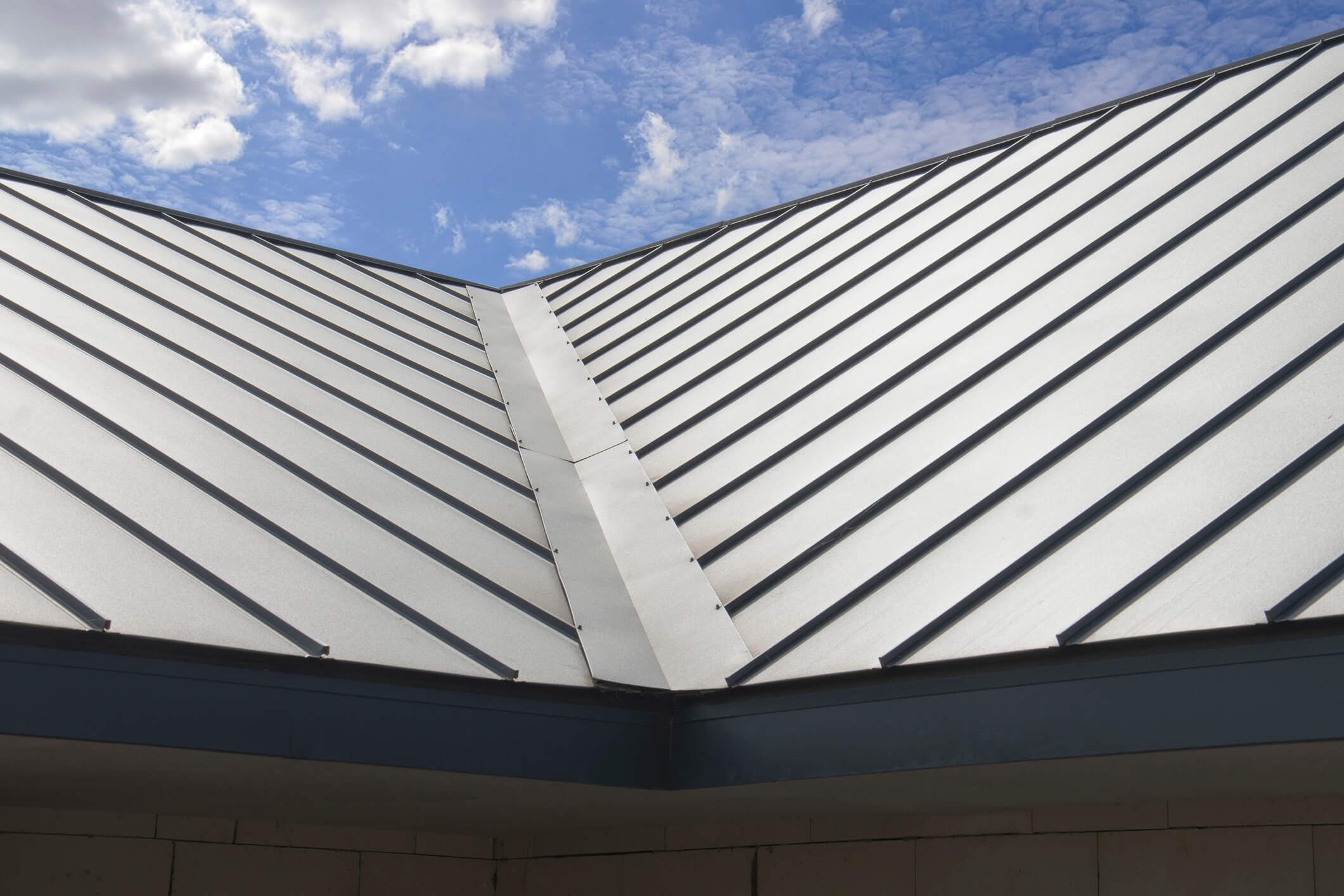 A close-up view of a modern metal roof with standing seam panels under a partly cloudy blue sky, showcasing expert craftsmanship inspired by New Orleans Shingle Roofing. The V-shaped valley highlights where two sections meet at an angle.