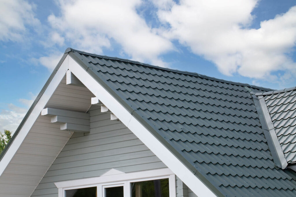 A close-up view of a house roof with gray metal roofing tiles, white trim, and light gray siding under a partly cloudy blue sky—showcasing quality metal roofing New Orleans homeowners can trust.