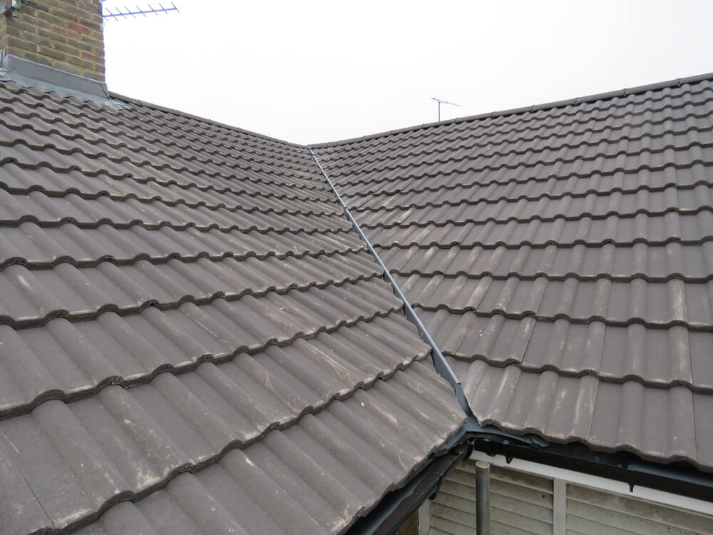 A close-up view of a tiled house roof showcases brown, wavy roof tiles and a prominent metal valley where two sections meet—an example of classic New Orleans Tile Roofing. Part of a chimney and antenna are also visible in the background.