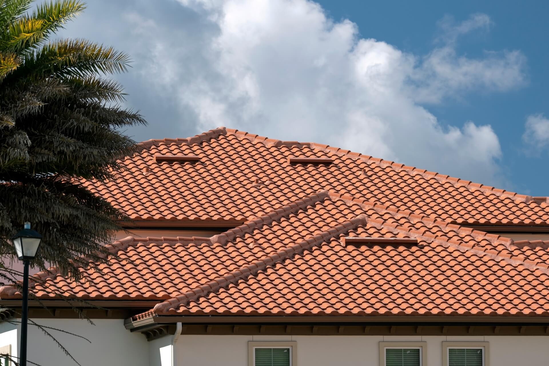 A close-up view of a house with multiple sloped roofs showcasing New Orleans Tile Roofing in reddish-orange clay tiles, set against a blue sky with fluffy white clouds and some tree branches on the left.