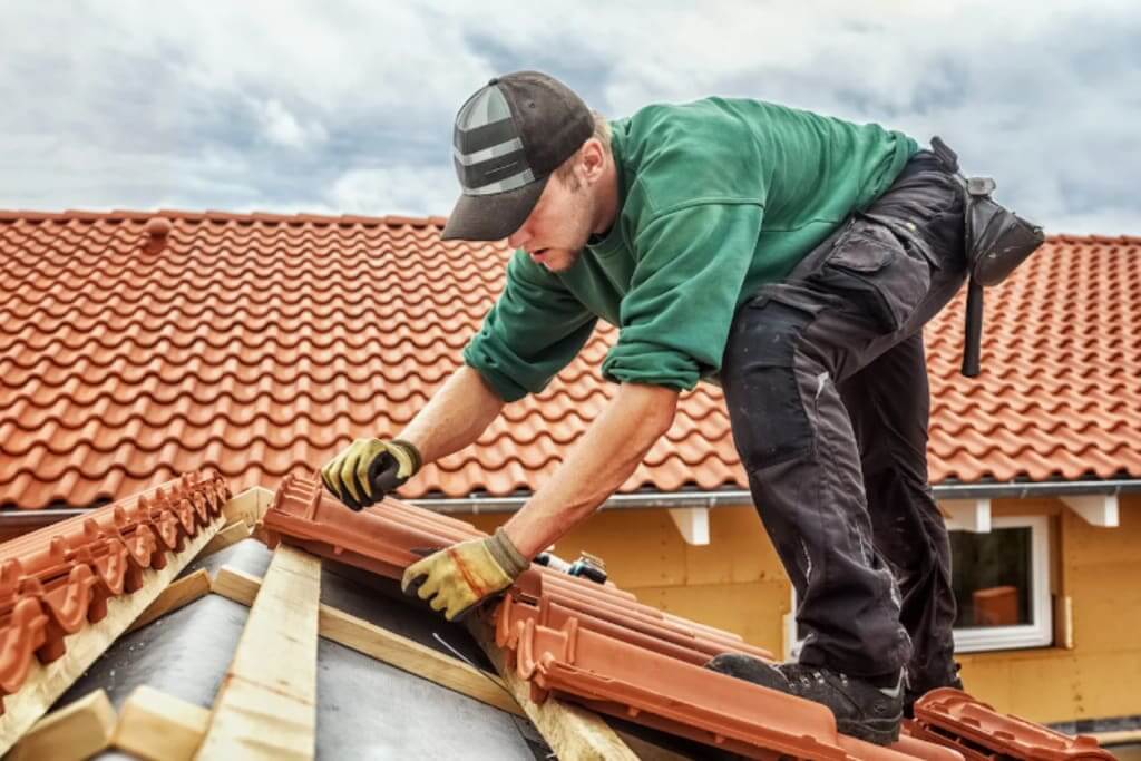 A worker in a green shirt and black cap installs red clay roof tiles on a house roof under a cloudy sky, using gloves and tools for the New Orleans Tile Roofing job. Another tiled roof and part of a house are visible in the background.