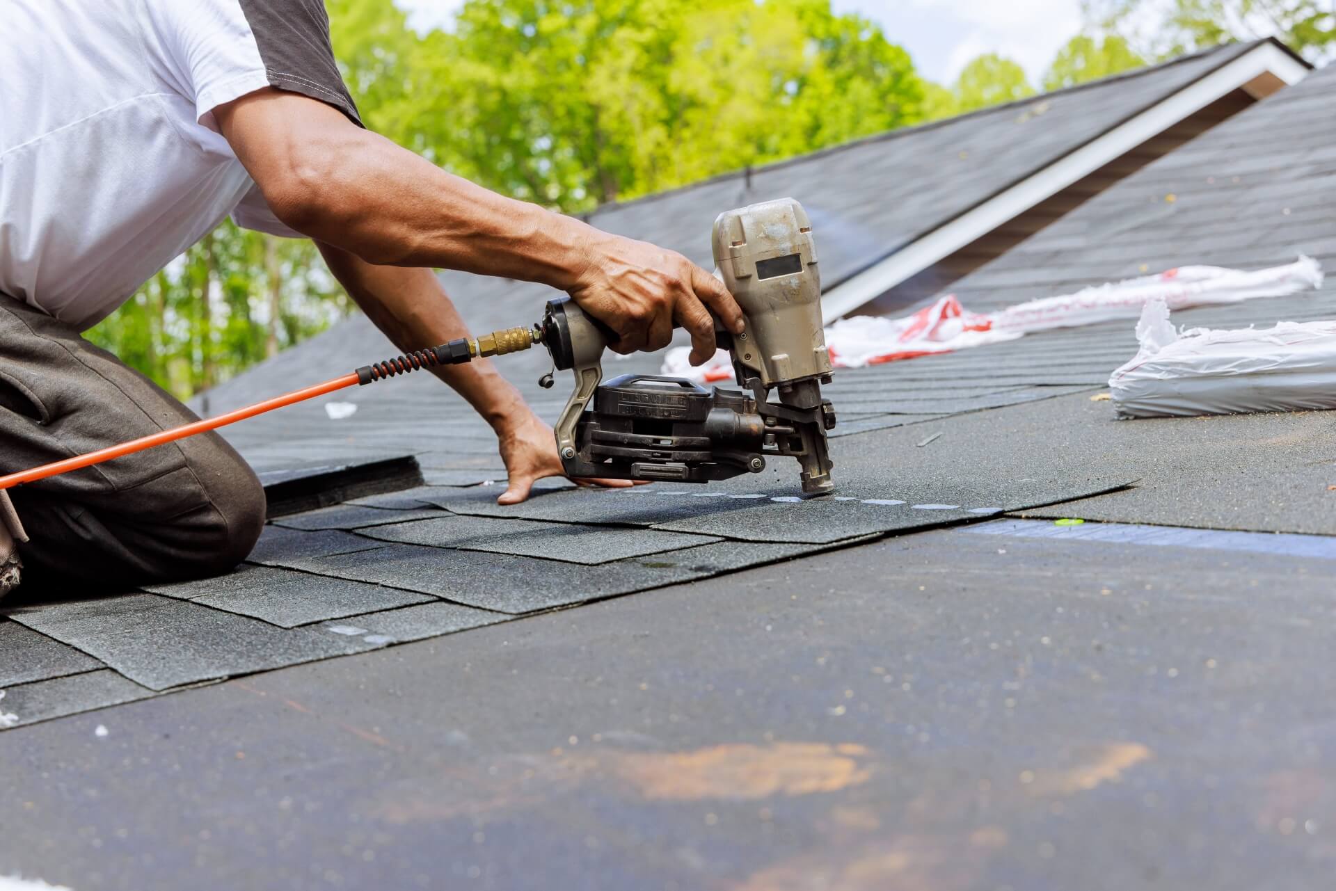 A person installs asphalt shingles on a roof using a nail gun, with bundles of roofing materials nearby and green trees in the background—showcasing expert Roof Repair New Orleans services.
