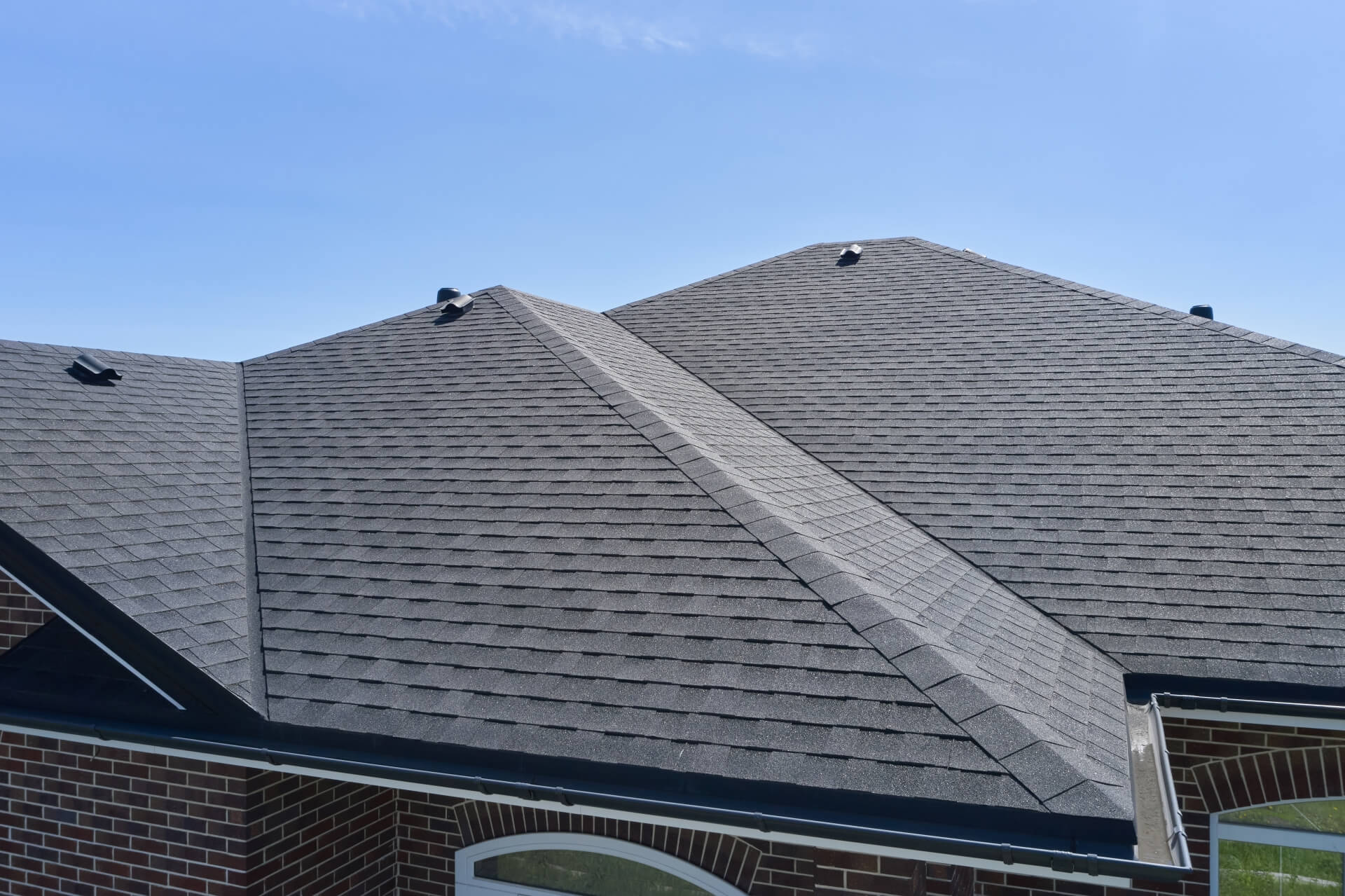 A close-up view of the rooftops of a brick house with gray asphalt shingles under a clear blue sky, perfect inspiration for anyone considering roof replacement in New Orleans. Multiple peaks, ridges, and roof vents are clearly visible.
