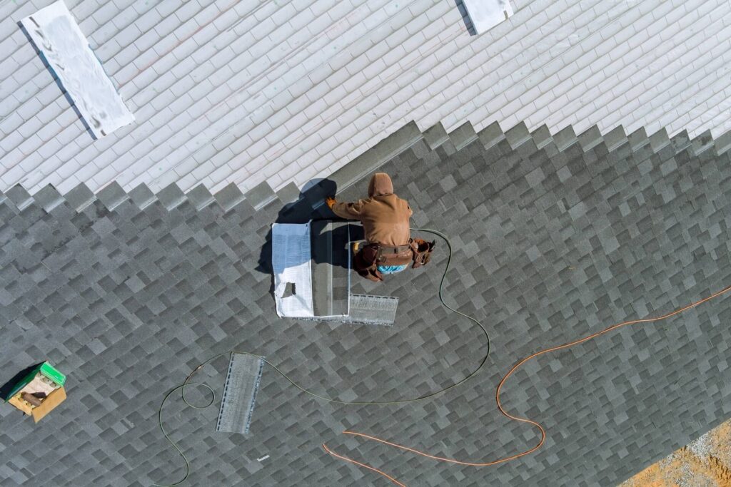 A person in a brown jacket and hood is diligently working on a grey shingled roof, surrounded by roof vents and tools. As part of a Roofing Company in New Orleans, they use equipment to install materials, with an orange extension cord and other items scattered around.