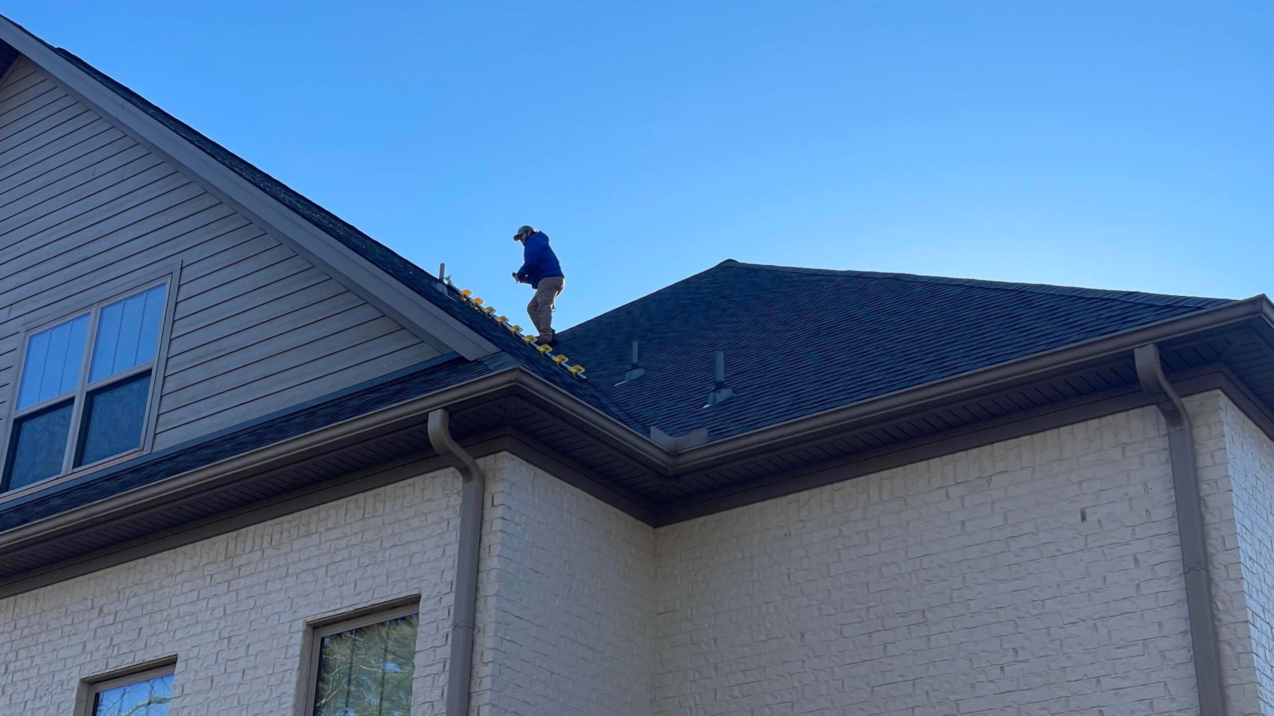 A person in a blue jacket and cap is on the roof of a two-story, light-brick house, clearing leaves from the gutter. The sky above New Orleans is clear and blue, making it an ideal day for any roofing company to check on your home's safety and efficiency.