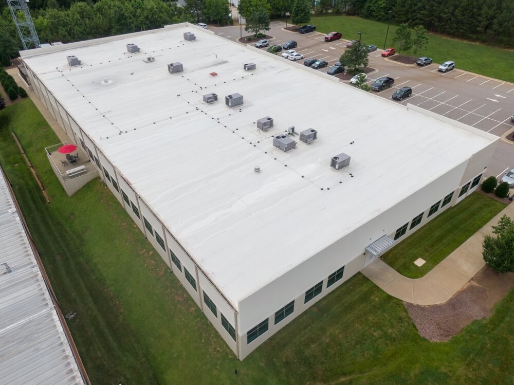 Aerial view of a large, rectangular, white-roofed warehouse crafted by a renowned roofing company in New Orleans, surrounded by lush green grass and trees. Several air conditioning units are on the roof. An adjacent parking lot with multiple cars is visible.