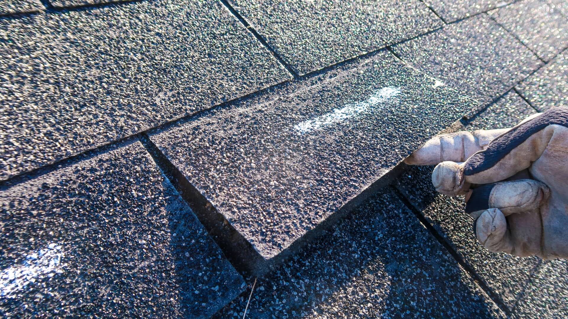 A gloved hand lifts an asphalt roof shingle on a rooftop, showing close-up detail of the textured shingles in sunlight—a common scene during Roofing Insurance Claims in New Orleans.