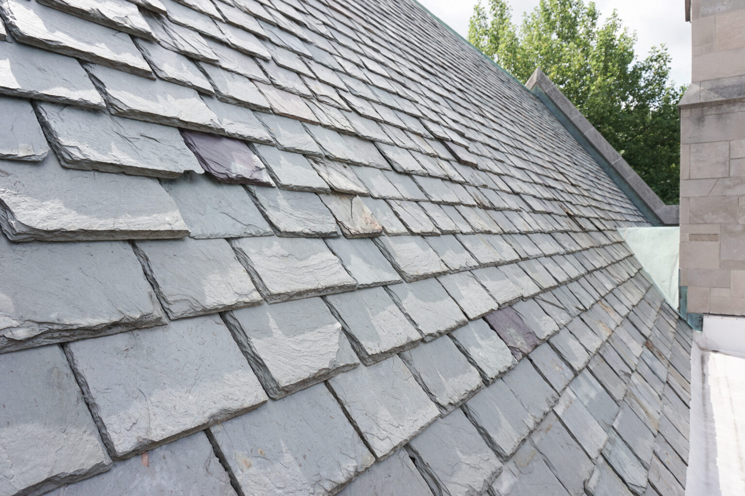 Close-up view of a sloped roof covered with overlapping gray slate tiles, showcasing classic Slate Roofing New Orleans style; some trees and part of a stone building appear in the background.